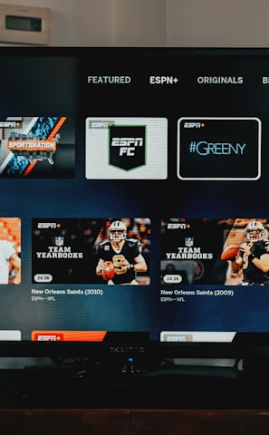 A television screen displaying the ESPN+ streaming interface with various sports-related shows and categories. The screen includes sections such as 'Browse by Shows' and 'Best of Drew Brees' with thumbnails of sports programs featuring athletes in action and analysts.